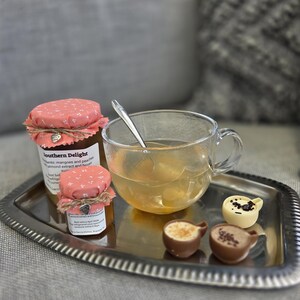 May include: A silver tray holds a glass teacup with a spoon, two small jars with pink floral lids, and three chocolate teacup-shaped sweets. The jars have labels, and the teacup contains a light-colored liquid. The background is blurred.
