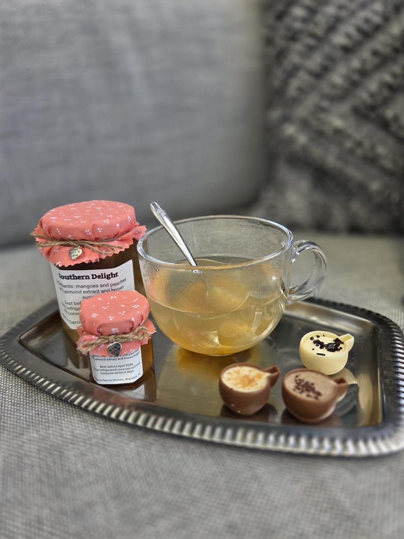 May include: A silver tray holds a glass teacup with a spoon, two small jars with pink floral lids, and three chocolate teacup-shaped sweets. The jars have labels, and the teacup contains a light-colored liquid. The background is blurred.