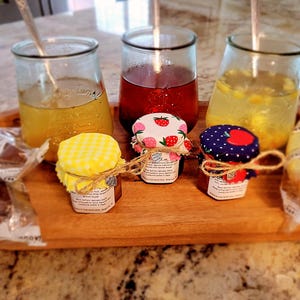 May include: A wooden tray displays three glass jars with drinks and three small jars of preserves. The preserve jars have fabric lids with strawberry and polka dot patterns. Wrapped breadsticks and spoons are also visible.