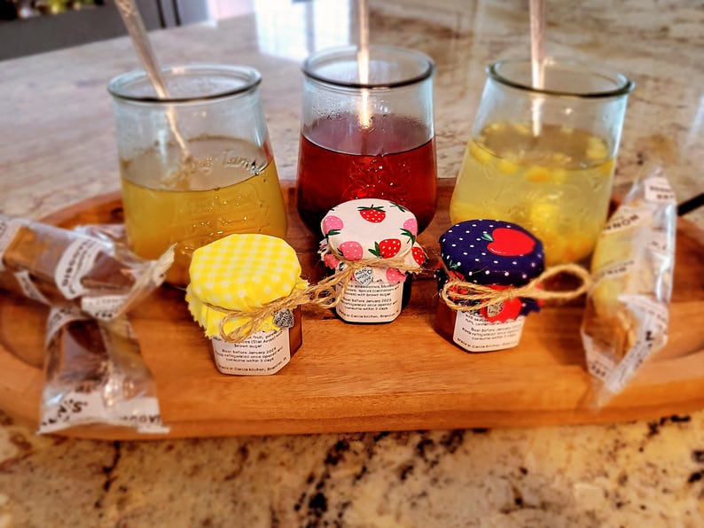 May include: A wooden tray displays three glass jars with drinks and three small jars of preserves. The preserve jars have fabric lids with strawberry and polka dot patterns. Wrapped breadsticks and spoons are also visible.