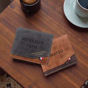 Peut inclure: Deux portefeuilles pliables sur une surface en bois. L'un est gris foncé avec "MEILLEUR PAPA" imprimé, l'autre est marron avec "MATEO" imprimé. Les deux ont un accent marron et un petit drapeau tricolore.