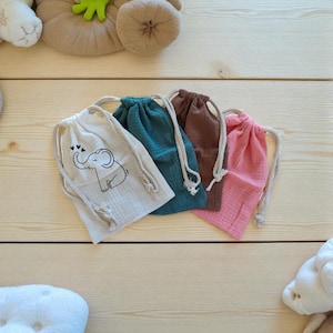 Peut inclure: Quatre sacs à cordon de serrage crème, turquoise, marron et rose, sur un fond en bois clair. Le sac crème présente un motif d'éléphant noir avec des cœurs. Chaque sac a une fermeture à cordon blanche.