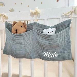 May include: A light green fabric crib organizer with two pockets, one holding a brown teddy bear and the other a white stuffed animal. The organizer has the name "Mylo" embroidered on the front.