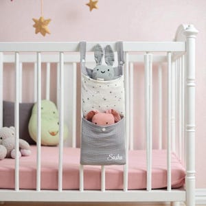 Può includere: Un organizer in tessuto grigio e bianco appeso a una culla bianca. L'organizer ha due tasche, una con un peluche a forma di coniglio grigio e l'altra con un peluche a forma di coniglio rosa. La tasca inferiore ha ricamato il nome "Sasha". La culla ha un materasso rosa.