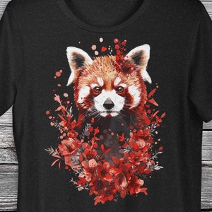 Puede incluir: Camiseta negra con una ilustración de un panda rojo rodeado de flores rojas. El panda tiene la cara blanca y los ojos negros.