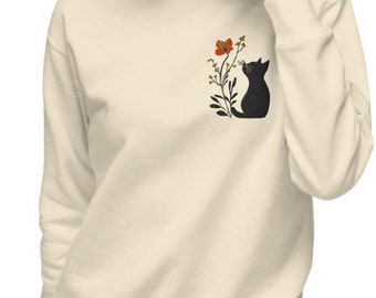 Op de afbeelding: Een crèmekleurig sweatshirt met een zwart silhouet van een kat die omhoog kijkt naar een rode bloem op een stengel met groene bladeren.