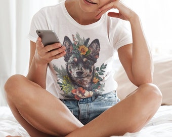 May include: White t-shirt featuring a dog portrait with a floral crown. The dog has a black and brown coat and is surrounded by colorful flowers. The person is sitting on a bed, wearing denim shorts, and holding a smartphone.