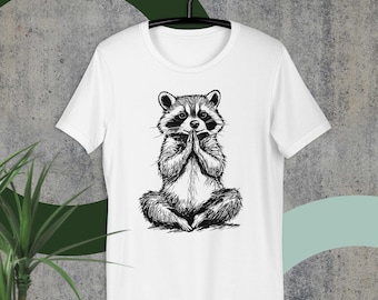 May include: White t-shirt featuring a black and white illustration of a raccoon sitting in a yoga pose with its hands in a prayer position.