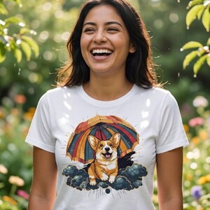 May include: White t-shirt with a cartoon corgi dog design sheltering under a rainbow umbrella. The dog is smiling. The person wearing the shirt is smiling. The background is a garden with flowers.