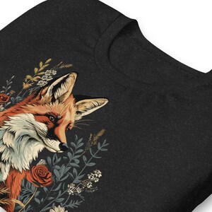 May include: Dark gray t-shirt featuring a detailed illustration of a fox with orange, white, and brown fur, surrounded by floral elements in shades of red, green, and cream. The design is on the left side of the shirt.
