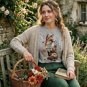 May include: A woman wearing a gray t-shirt with a fox graphic, a beige cardigan, and green pants, sitting on a wooden bench. A wicker basket filled with flowers and a book are in her lap. The scene is set in a garden with a stone building in the background.