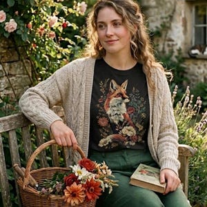 May include: A woman wearing a black t-shirt with a fox illustration, a beige cardigan, and green pants, sitting on a wooden bench. A wicker basket filled with flowers and a book are on the bench. The scene is set outdoors with a stone building and flowers.
