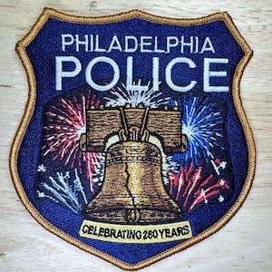 May include: A blue and gold embroidered patch featuring the words "PHILADELPHIA POLICE" above an image of the Liberty Bell. Red, white, and blue fireworks burst behind the bell. The text "CELEBRATING 250 YEARS" is at the bottom.