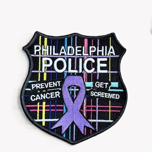 May include: A black and white embroidered patch with the words "Philadelphia Police" in white. The patch features a purple ribbon with the words "Prevent Cancer" and "Get Screened" on either side. The background of the patch is a colorful plaid pattern.