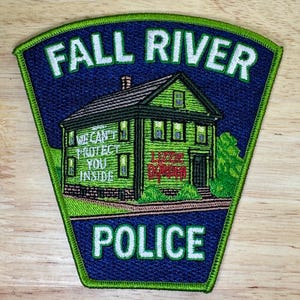 May include: A Fall River Police patch with a green and blue design. The patch features a green house with the text "Lizzie Borden" and "We Can't Protect You Inside". The words "Fall River Police" are at the top.
