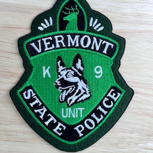 May include: Green and black embroidered patch with a white German Shepherd dog head, the text "K9" above the dog, "UNIT" below, and the text "VERMONT STATE POLICE" around the outside.