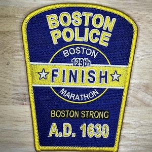 May include: A blue and yellow embroidered patch with the words "Boston Police" and "Boston Strong" and the text "A.D. 1630". The patch also features a circle with the words "Boston 129th Marathon Finish" and two stars.