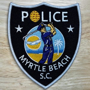 May include: Embroidered police patch featuring a shield design with the word "POLICE" at the top. The patch includes a golf scene with a police officer swinging a golf club. The text "MYRTLE BEACH S.C." is at the bottom.
