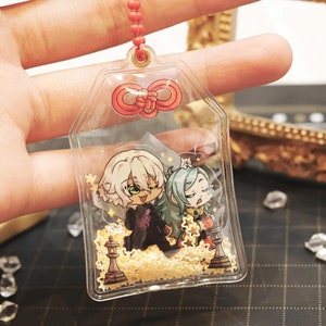 May include: A clear plastic keychain charm with a red bow on top. Inside the charm are two anime characters, one with white hair and the other with green hair, standing on a bed of gold glitter.