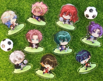 May include: A collection of acrylic charms featuring anime-style characters in soccer uniforms. Each charm has a unique character design with different hair colors and outfits. Two soccer balls are also present on a green, grassy surface.