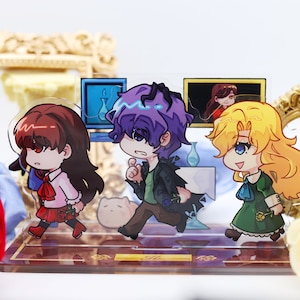 May include: A set of three acrylic character stands featuring anime-style characters. The characters are a girl with brown hair and a red dress, a boy with purple hair and a black jacket, and a girl with blonde hair and a green dress. The stands are on a brown base with a gold trim.