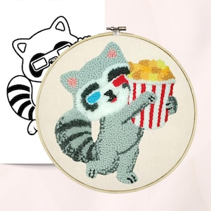 May include: A punch needle PDF pattern featuring a raccoon wearing 3D glasses and holding a popcorn bucket. The raccoon is gray, white, and black, with a red and white striped popcorn bucket. The text "Punch Needle PDF Pattern" is at the top.