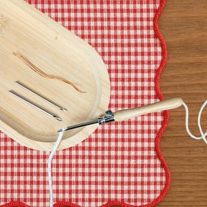Pode incluir: Uma ferramenta de agulha mágica de madeira clara e agulha de metal, posicionada sobre fio branco. A ferramenta está sobre tecido xadrez vermelho e branco com borda ondulada, ao lado de uma bandeja de madeira com agulhas e fio.