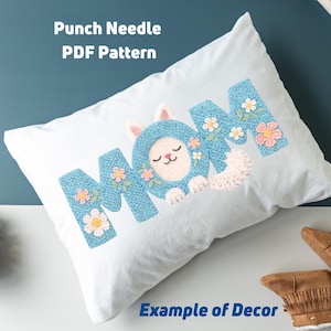 May include: White pillow with the word "MOM" embroidered in blue yarn, featuring a cat design in the letter "O". The letters are decorated with floral accents. Text on the pillow reads "Punch Needle PDF Pattern" and "Example of Decor".