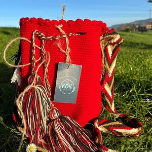 May include: Red crocheted bag with a long, colorful strap and tassels. The bag has a label that says "KOHE - the art of style".