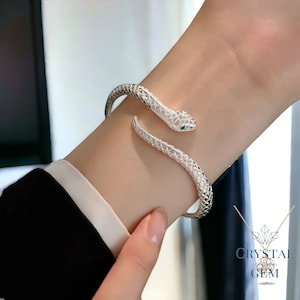 May include: A silver snake-shaped cuff bracelet. The bracelet wraps around the wrist, with the snake's head and tail meeting. The snake's head features a small green gem for the eye, and the body is encrusted with small, clear gems.