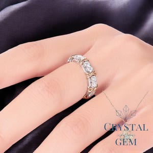 May include: A silver and gold ring with clear gemstones. The ring features a repeating pattern of three gemstones, with gold accents and a silver band. The ring is on a person's finger, with a dark background.