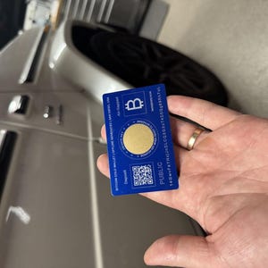 May include: A blue plastic card with a gold circle in the center. The card has the text "Bitcoin Cold Wallet | Offline Generated | Made in America" and "PUBLIC 146WVFAYNJ43J4C605X7AC1010588R71UL" printed on it. The card also has a QR code and the text "Deposit" printed on it.
