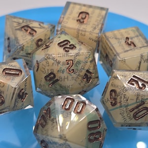 May include: A set of seven polyhedral dice with a vintage map design encased in clear resin. The dice have brown numbers and are arranged on a blue plate, ideal for tabletop games.