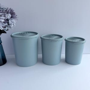 May include: A set of three cylindrical storage containers in a muted dusty blue color. Each container features a matching circular lid with a sunburst pattern radiating from the center. The containers are arranged in descending order of size.