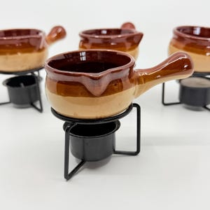 May include: Four small ceramic pots with handles, each featuring a two-tone brown and tan glazed finish. Each pot rests on a black metal wire stand designed to hold a small heat source underneath the bowl for warming contents during service.