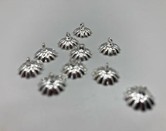 May include: A collection of silver-coloured, flower-shaped jewellery findings. Each finding has a small loop at the top and a scalloped edge. The findings are scattered on a white surface.