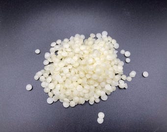 May include: A collection of small, off-white wax beads on a dark grey surface. The beads are round and slightly irregular, with a matte finish. The wax beads are scattered across the surface.