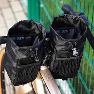 Puede incluir: Dos bolsas de manillar negras con bolsillos de malla y cierre de cordón. Las bolsas están sujetas al manillar de una bicicleta y tienen el texto "X-Pac" impreso en ellas.