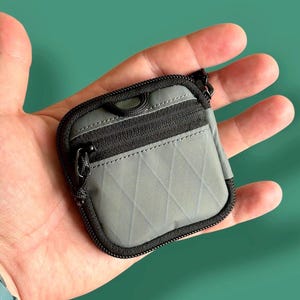 Puede incluir: Pequeño estuche gris con cremallera y cremallera y ribete negros. El estuche se sostiene en una mano y tiene un patrón de diamantes en la parte delantera. El estuche tiene un pequeño lazo en la parte superior y un pequeño tirador.