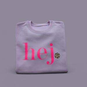 Könnte beinhalten: Ein lavendelfarbener Pullover mit dem Wort "hej" in Pink auf der Vorderseite. Neben dem Wort befindet sich ein kleines Leopardenmuster.