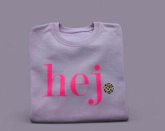 Puede incluir: Una sudadera de cuello redondo en color morado claro con la palabra hej. impresa en un tono rosa neón brillante en el pecho, con un pequeño círculo de estampado de leopardo que sustituye al punto sobre la letra j de hej.