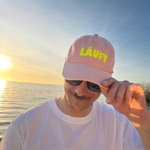 Könnte beinhalten: Hellrosa Baseballkappe mit dem Wort "LÄUFT" in leuchtend gelben Blockbuchstaben. Die Kappe wird von einer Person getragen, die eine Sonnenbrille und ein weißes T-Shirt trägt, mit einem Sonnenuntergang und Wasser im Hintergrund.