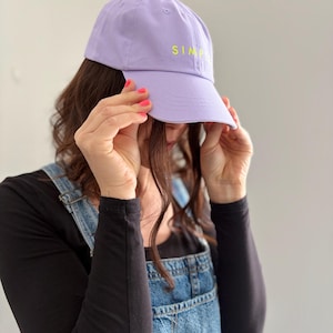 Op de afbeelding: Een lila baseballpet met het woord "SIMP" in geel geborduurd. De pet is gemaakt van een zachte stof en heeft een gebogen klep. De persoon draagt een zwart shirt met lange mouwen en een spijkerbroek met tuinbroek.