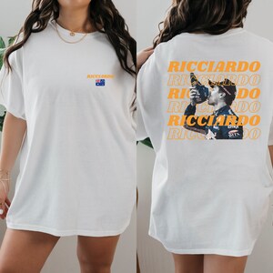 May include: Two white t-shirts featuring a graphic print of Formula One driver Daniel Ricciardo. The front of one shirt has the word "Ricciardo" printed in orange. The back of the other shirt has the word "Ricciardo" printed in orange multiple times with a picture of Ricciardo celebrating.