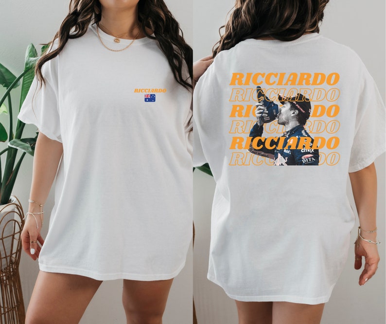 May include: Two white t-shirts featuring a graphic print of Formula One driver Daniel Ricciardo. The front of one shirt has the word "Ricciardo" printed in orange. The back of the other shirt has the word "Ricciardo" printed in orange multiple times with a picture of Ricciardo celebrating.
