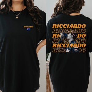 May include: Black t-shirt with a graphic of Formula One driver Daniel Ricciardo on the back. The graphic is repeated multiple times in orange text. The front of the shirt has a small Australian flag and the text "Ricciardo" in orange.