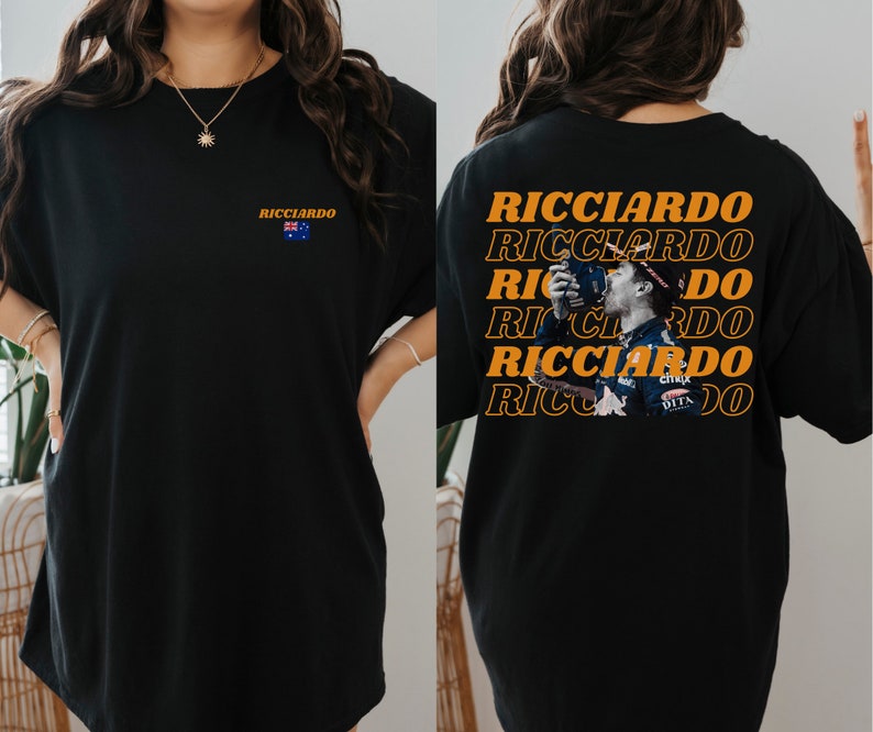 May include: Black t-shirt with a graphic of Formula One driver Daniel Ricciardo on the back. The graphic is repeated multiple times in orange text. The front of the shirt has a small Australian flag and the text "Ricciardo" in orange.