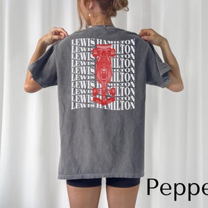 Peut inclure: Un t-shirt gris avec un motif rouge et blanc d'une voiture de course au dos. Le texte "LEWIS HAMILTON" est répété plusieurs fois sur le t-shirt.
