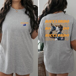 May include: Grey t-shirt featuring a graphic of a racing driver drinking from a bottle. The graphic is repeated several times and the text "RICCIARDO" is printed in orange. The shirt also has a small Australian flag printed on the front.