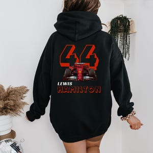 Może przedstawiać: Czarna bluza z kapturem z grafiką czerwonego i białego samochodu wyścigowego i napisem "44 LEWIS HAMILTON" nadrukowanym na plecach.
